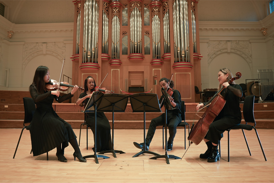 Four students, wearing formal attire, performing various string instruments, seated in a semi-circle facing the audience.